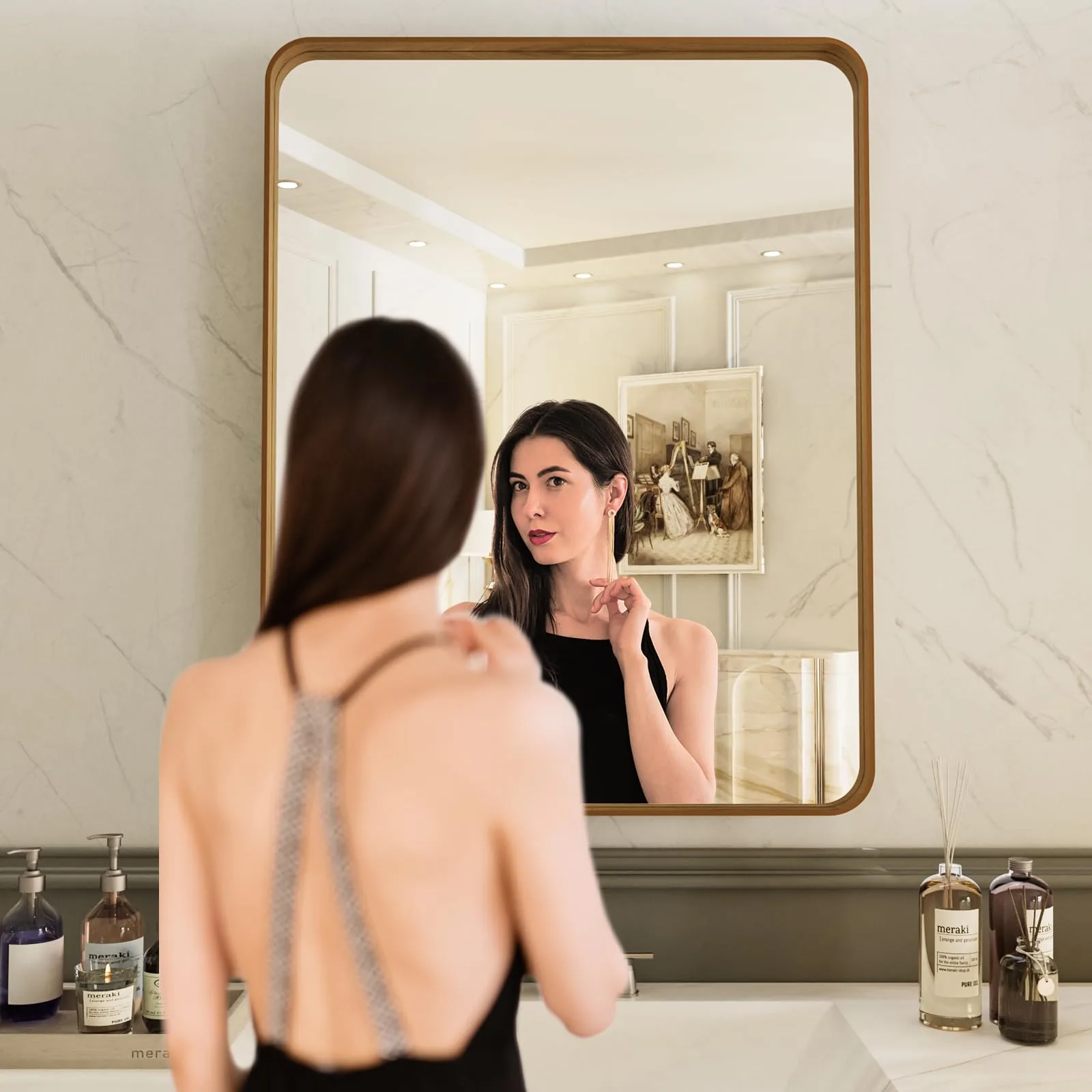 A woman checking her reflection in a gold-framed rounded rectangle mirror above a marble bathroom vanity